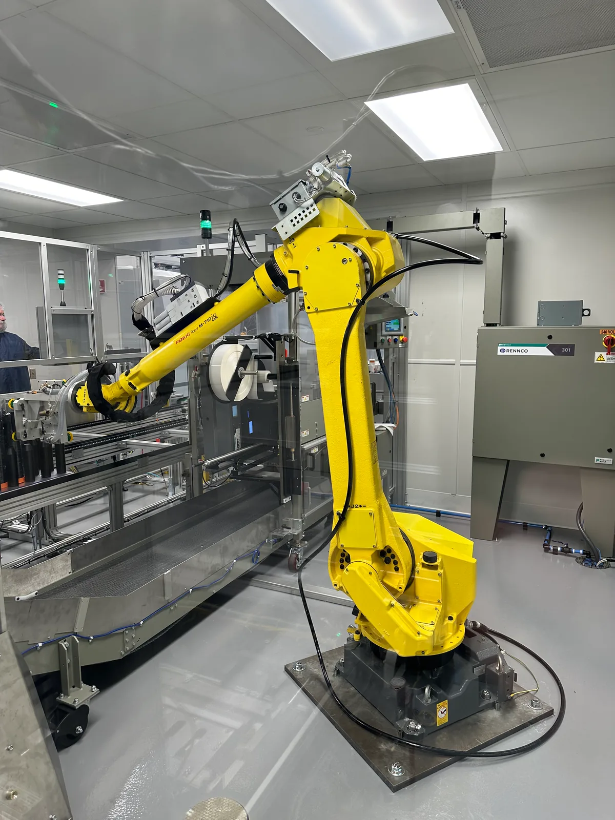 A FANUC robotic arm integrated with a Rennco packaging machine and stainless infeed conveyor in a cleanroom packaging cell.